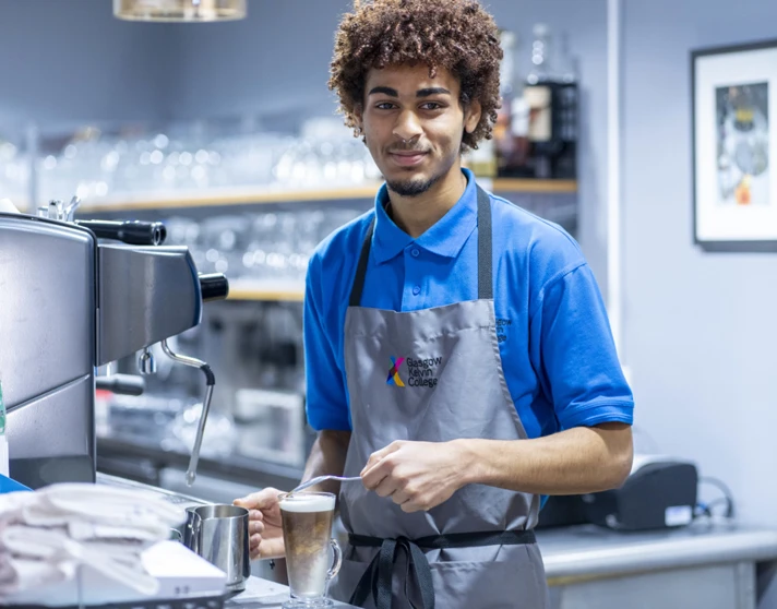 A young male barista in a blue uniform and grey apron, smiling while preparing a latte at a coffee machine in a café setting. A young male barista in a blue uniform and grey apron, smiling while preparing a latte at a coffee machine in a café setting.
